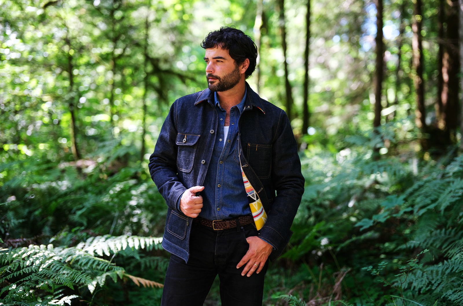 A man wearing a dark denim jacket over a blue button-down shirt stands in a lush green forest with ferns.